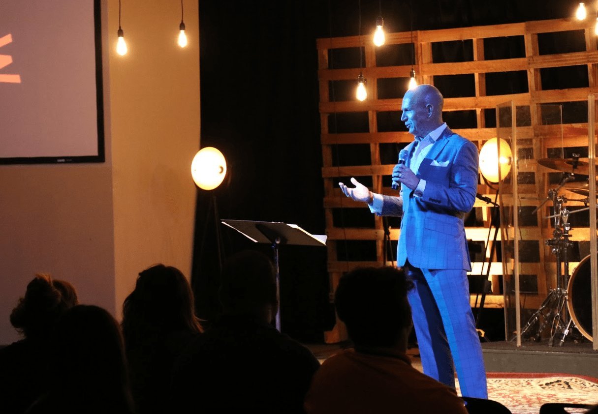 Man in blue suit speaking at podium on stage with audience silhouettes in foreground and shelving with books in background
