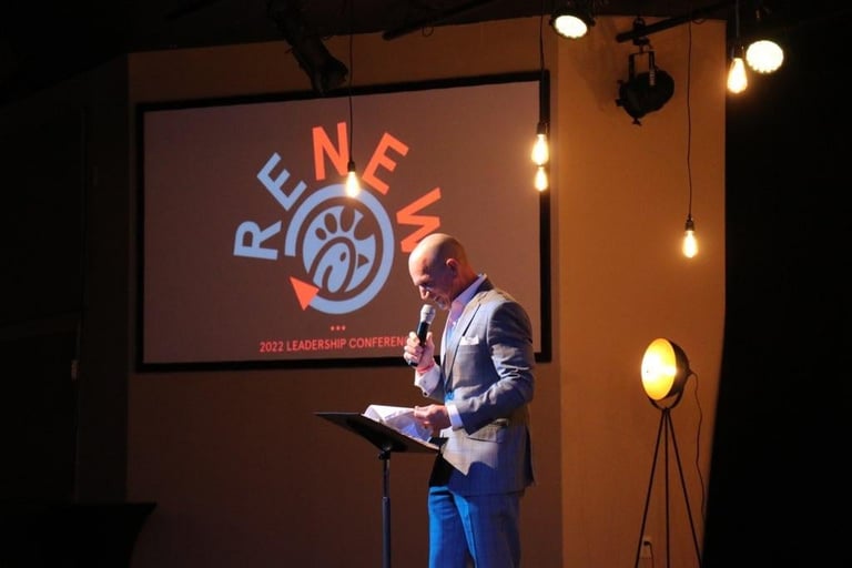 Man in blazer speaking at podium during 2024 Leadership Conference with Renew logo on screen behind him