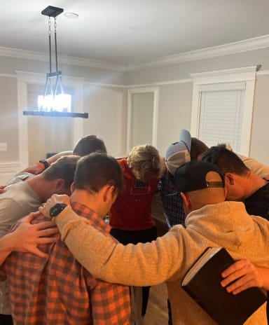 Group of people standing in a circle with arms around each other in a home interior, wearing orange and red clothing