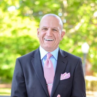 Smiling older man in dark suit with pink tie and pocket square against blurred green foliage background