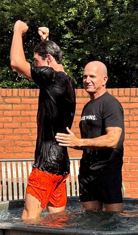 Two men in a pool, one wearing a black wet shirt with fist raised in celebration, the other in black shirt smiling beside him