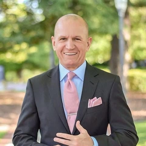 Professional headshot of smiling middle-aged man in black blazer, light blue shirt, and pink tie outdoors with blurred green trees background